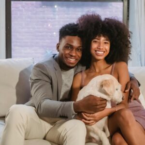 Happy young Black couple sitting on comfortable sofa with cushions and stroking adorable dog while spending time together in living room