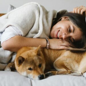 Cheerful brunette in comfy wear embracing fluffy adorable Shiba Inu dog while lying together on cozy couch in light living room